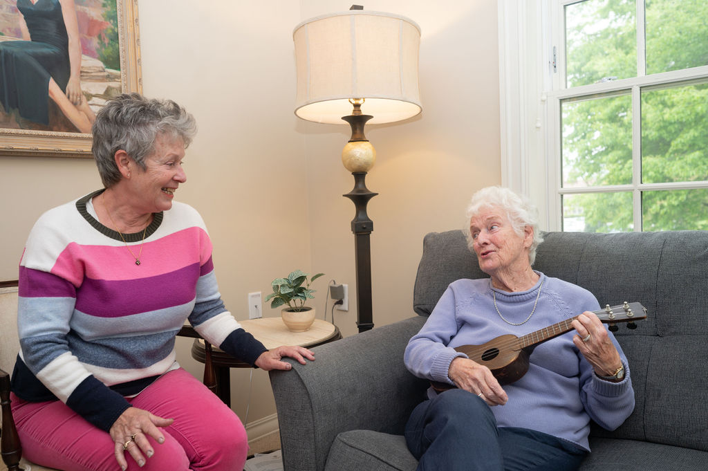 Two residents are seated, chatting. One holds a ukelele as they enjoy the community life at Hearth at Drexel